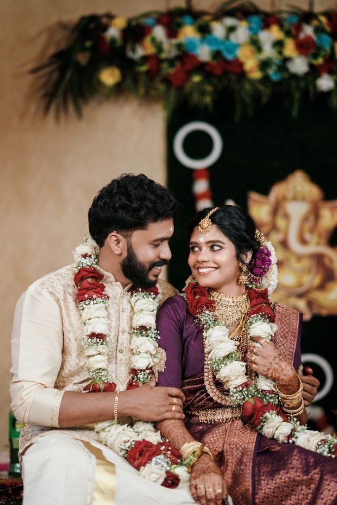 Traditional Indian wedding couple in purple and gold attire with floral garlands and henna, seated before Lord Ganesha decor – Al Ameen Wedding Planner Kochi.