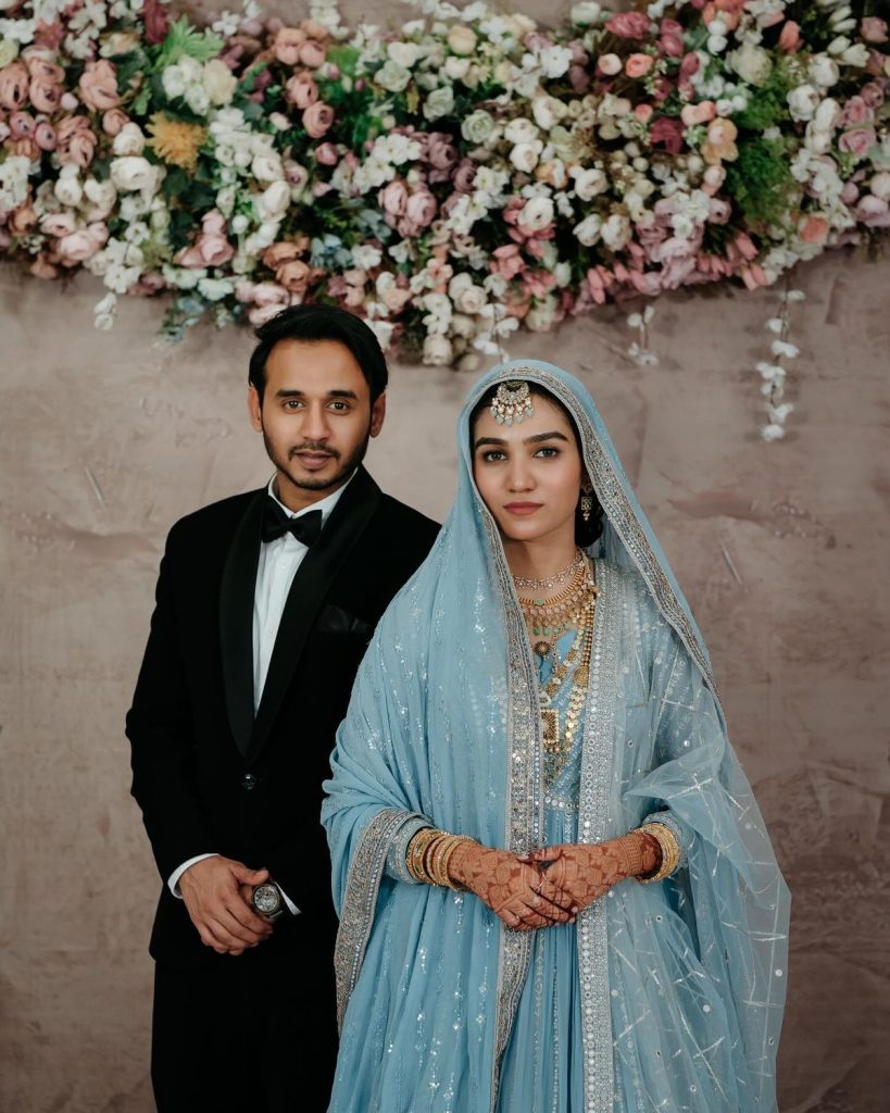 Traditional Indian bride in ornate blue embroidered attire with henna and jewelry, standing beside groom in black tuxedo during luxury Kerala wedding ceremony — floral backdrop by Al Ameen Wedding Planner, Kochi.