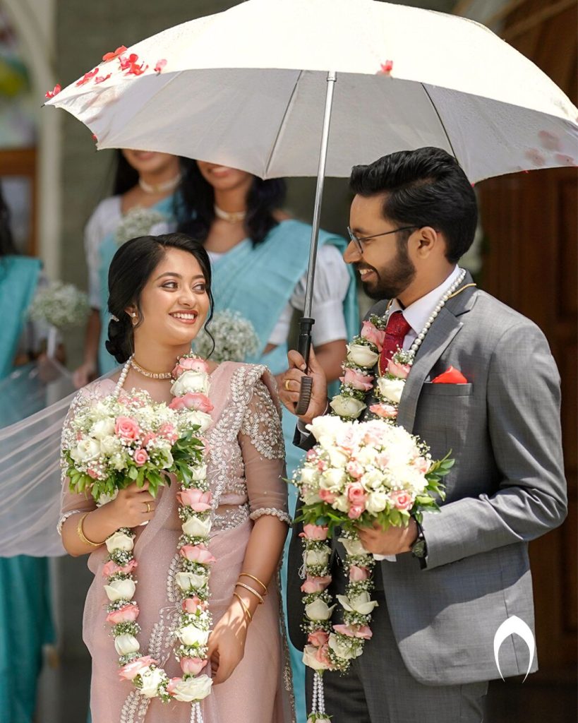 Bride and groom in traditional Indian wedding attire with floral garlands, standing under a white umbrella, surrounded by bridesmaids in blue sarees — outdoor ceremony captured by Al Ameen Wedding Planner, Kochi.